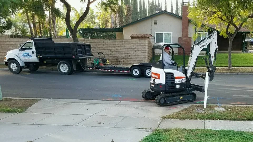Mini excavator on driveway with dump truck and flatbed trailer for underground plumbing access