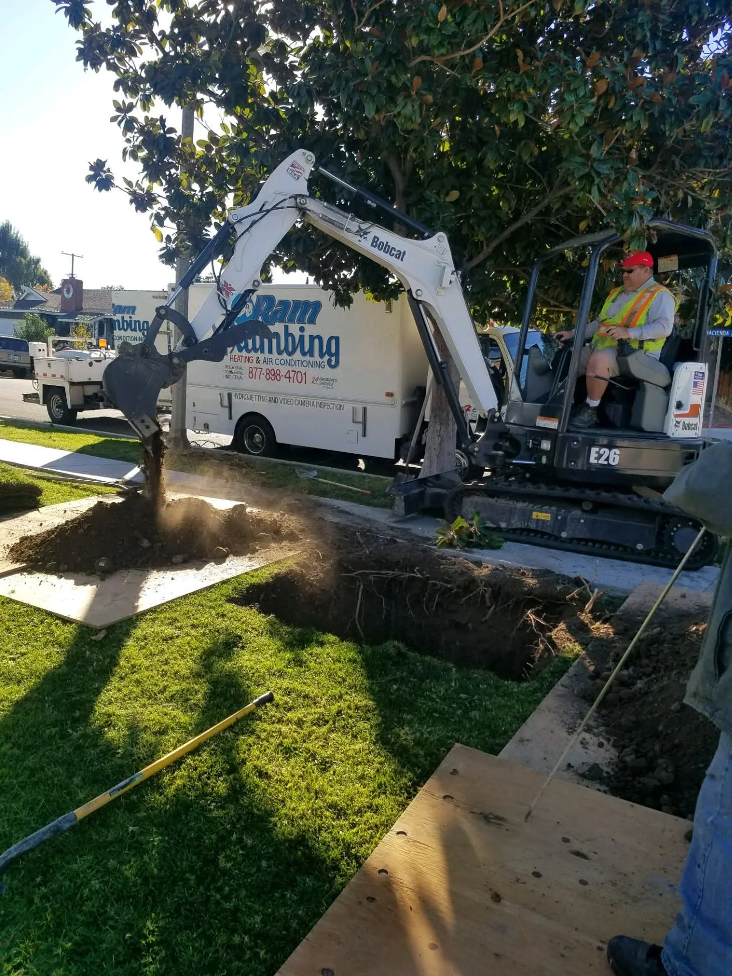 Mini excavator digging sewer line trench through residential lawn with mature trees