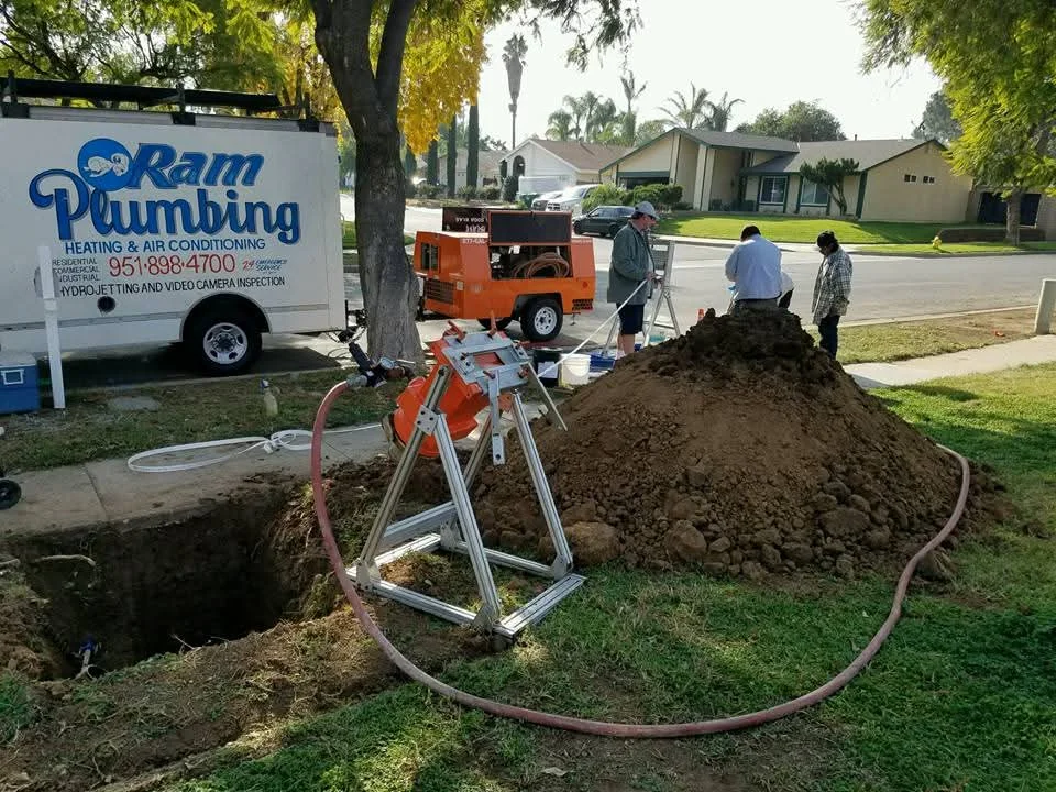 Sewer line excavation with soil pile, auger equipment, and hydro-jetting hose on residential property